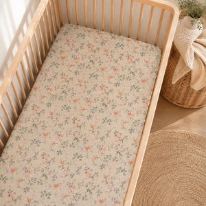 Puede incluir: Una sábana rectangular para cuna con fondo beige claro y un patrón repetitivo de pequeñas flores rosas y follaje verde suave. La tela está ajustada sobre un marco de colchón de madera clara con barrotes verticales.