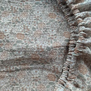May include: Close-up of a floral patterned fabric in shades of beige, brown, and green. The fabric has a delicate, vintage aesthetic, with a ruffled edge on the right side. The pattern features small flowers and leaves.