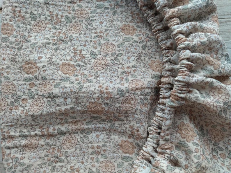 May include: Close-up of a floral patterned fabric in shades of beige, brown, and green. The fabric has a delicate, vintage aesthetic, with a ruffled edge on the right side. The pattern features small flowers and leaves.