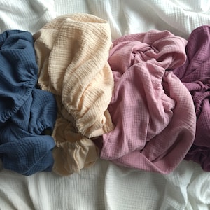 May include: Four pieces of crinkled muslin fabric in shades of dark blue, cream, dusty rose, and deep plum. Each piece features a gathered elastic edge, creating a soft, textured appearance with a lightweight, layered, and breathable cotton weave.