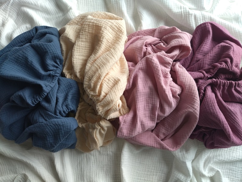 May include: Four pieces of crinkled muslin fabric in shades of dark blue, cream, dusty rose, and deep plum. Each piece features a gathered elastic edge, creating a soft, textured appearance with a lightweight, layered, and breathable cotton weave.