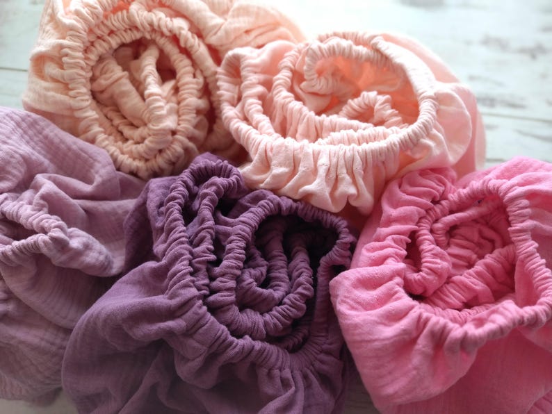 May include: A collection of five gathered fabric pieces in various shades of pink and purple. The textiles feature a crinkled texture and elasticized edges, creating a soft, layered appearance with colors ranging from pale peach to deep magenta.