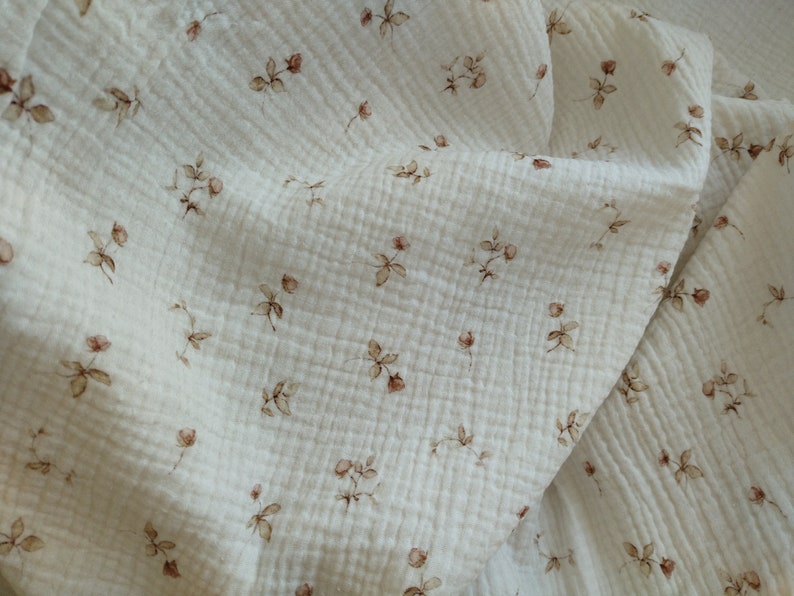 May include: Off-white muslin fabric with a delicate floral pattern. The fabric features small, brown rose illustrations scattered across its surface. The material has a textured, crinkled appearance, suggesting a soft and breathable quality.