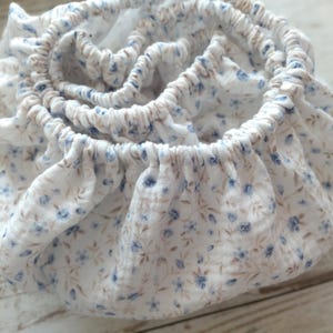 May include: A white fabric item with an elasticized edge, patterned with small blue and tan floral designs. The fabric is gathered, creating a ruffled effect. The item appears to be a fitted sheet or similar bedding accessory.