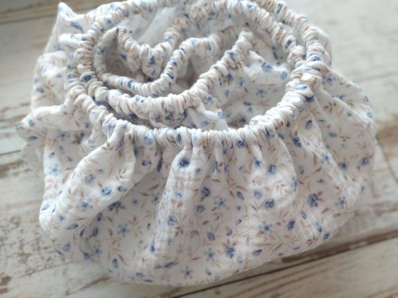 May include: A white fabric item with an elasticized edge, patterned with small blue and tan floral designs. The fabric is gathered, creating a ruffled effect. The item appears to be a fitted sheet or similar bedding accessory.