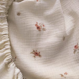 May include: Close-up of a cream-colored fitted sheet with a delicate floral pattern. The fabric has a textured appearance, and the elastic edge is visible on the left side. The floral design features small clusters of pink and brown flowers.