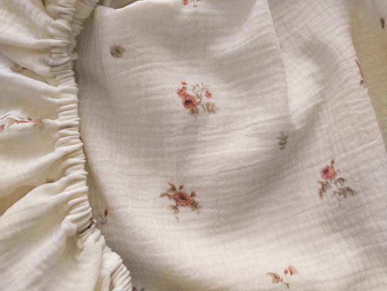 May include: Close-up of a cream-colored fitted sheet with a delicate floral pattern. The fabric has a textured appearance, and the elastic edge is visible on the left side. The floral design features small clusters of pink and brown flowers.
