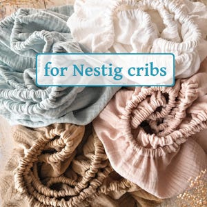 May include: A collection of four fitted crib sheets in white, light blue, light pink, and tan, each featuring an elasticized edge. The fabric has a textured, crinkled appearance. A rectangular label with a blue border displays the text for Nestig cribs.