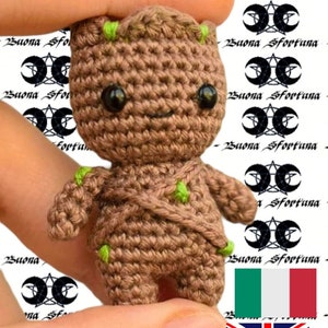 Pode incluir: Um pequeno personagem Groot de crochê do filme Guardiões da Galáxia. O personagem é marrom com detalhes verdes e tem uma expressão amigável. O personagem está sendo segurado em uma mão.