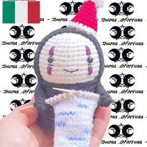 May include: A white crocheted No Face character from the movie Spirited Away wearing a red Santa hat and knitting a blue and white striped scarf.
