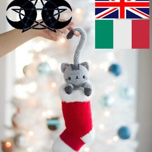 Puede incluir: Una media de Navidad roja y blanca con un gato de crochet gris asomándose por la parte superior. La media está colgada de un árbol de Navidad blanco.