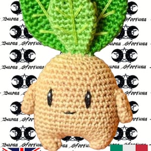 Può includere: Un giocattolo di peluche all'uncinetto a forma di rapa con un cappello verde a foglie e un viso sorridente. Il giocattolo è in piedi su uno sfondo bianco con un motivo ripetuto di mezze lune nere e stelle.