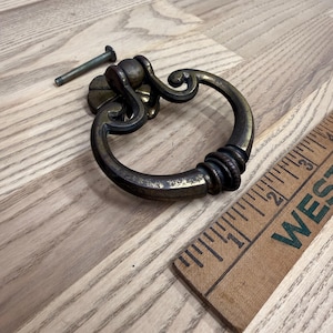 May include: An antique brass ring pull handle with a decorative backplate and a matching metal bolt. A wooden ruler is positioned alongside the hardware, showing the ring pull measures approximately 3 inches in total width and height.
