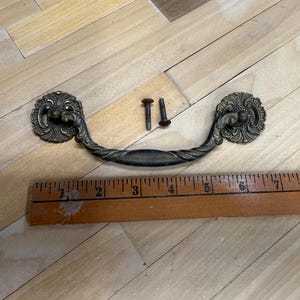 May include: An antique brass drawer pull with ornate floral detailing. The pull is curved with a central handle and two decorative end pieces. Two screws are included. A wooden ruler is in the background.