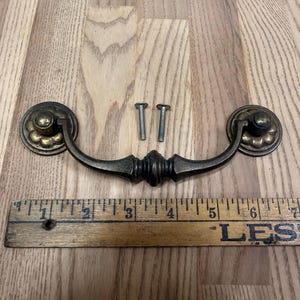 May include: A dark bronze-toned metal drawer pull with a curved handle and two circular backplates featuring a scalloped edge design. Two matching metal screws are included. A wooden ruler is visible, showing the handle width is 5 inches.