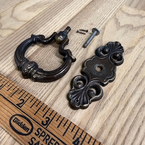 May include: A dark bronze-toned metal drop pull handle with a circular ring and a matching decorative backplate. The set includes two small screws and one bolt. A wooden ruler is visible, showing the hardware measures approximately 4 inches.