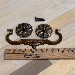 May include: Antique brass drawer pulls and matching rosettes. The drawer pull is 6 inches long. The rosettes are flower-shaped with a central knob. A screw is included. The hardware is a warm, aged brass color.
