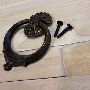 May include: An antique bronze-toned drawer pull with an oval ring and decorative backplate. Two black screws are included for installation. The hardware has a vintage aesthetic, suitable for furniture restoration or decorative projects.