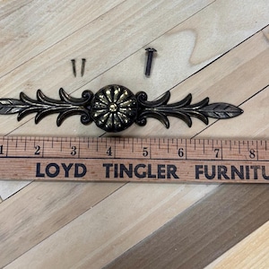 May include: A gold-toned metal drawer pull with a floral design. The pull has two leaf-shaped ends and a central round knob with a raised, sunburst pattern. Two screws are included.