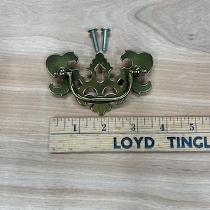 May include: A gold-toned metal drawer pull with an ornate design. The pull has a D-shaped handle and two screws for installation.