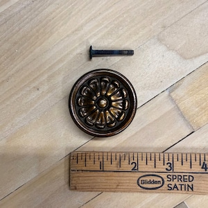 May include: A bronze-colored, round cabinet knob with a floral design. A dark screw is positioned above the knob. A wooden ruler is placed below the knob, showing inch measurements. The knob has a vintage aesthetic.