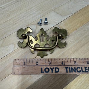 May include: A gold-toned metal cabinet pull with an ornate design. The pull has a decorative handle and two screws for installation.