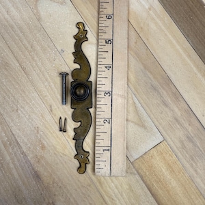 May include: An antique brass-toned cabinet pull with ornate scrollwork, a central knob, and mounting hardware. The pull measures approximately 6 inches in length. A wooden ruler is placed next to the pull for scale, showing the measurements in inches.