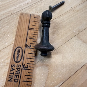 May include: A dark bronze-colored cabinet knob with a decorative top and a screw. The knob is approximately 3 inches tall. A wooden ruler is next to the knob.