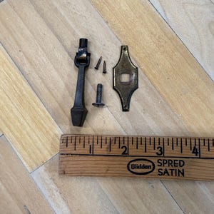 May include: An assortment of antique brass hardware components, including a handle, a decorative plate, and screws. A wooden ruler with the text "Bliddan SPRED SATIN" is also present, providing scale. The hardware appears aged and may be for furniture restoration.