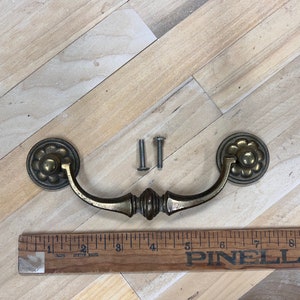 May include: Antique brass drawer pull with decorative floral accents and a curved handle. Includes two silver screws for installation. A wooden ruler is also in the image.