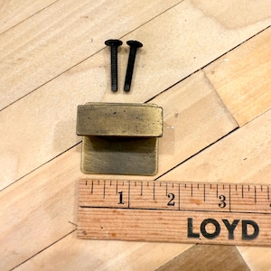 May include: Aged brass-toned cabinet hardware set, including a rectangular latch, two black screws, and a wooden ruler for scale. The latch is approximately 1 inch long. The hardware is set against a light wood grain surface.