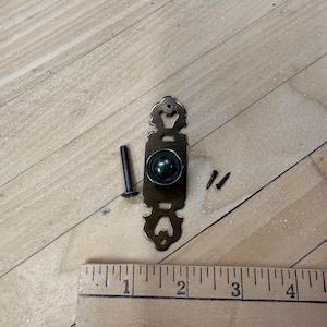 May include: An antique brass-toned cabinet pull with a round center and decorative cutouts. Includes a long screw and two smaller screws. A wooden ruler is placed next to the hardware, showing measurements in inches.