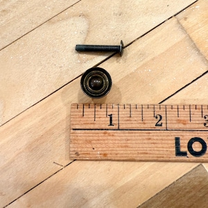 May include: A vintage brass cabinet knob with a matching screw, shown with a wooden ruler. The knob is round with a dark, aged finish. The ruler shows measurements in inches.