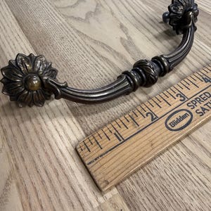 May include: A dark bronze-toned metal drawer pull featuring a curved handle with decorative floral-shaped mounting plates at each end. A wooden ruler is positioned alongside the handle, indicating a total length of approximately 4 inches.