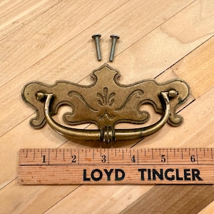 May include: Antique brass drawer pull with decorative flourishes and a curved handle. Includes two screws for installation. A wooden ruler with the text "LOYD TINGLER FURN" is placed below the drawer pull. The drawer pull measures approximately 6 inches in length.