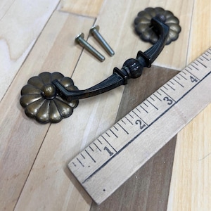 May include: A brass cabinet pull with a decorative flower design on each end. The pull is approximately 2 inches long and comes with two screws.