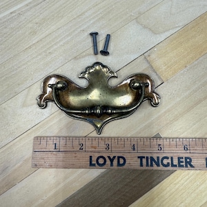 May include: Antique brass drawer pull with two screws. The pull has a decorative design with a curved handle and a central, ornate plate. A wooden ruler with the text "LOYD TINGLER" is also present.