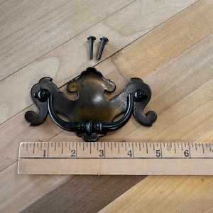 May include: Antique bronze-toned drawer pull with a decorative design, featuring a curved handle and ornate backplate. Includes two screws for installation. A wooden ruler is placed below the drawer pull.