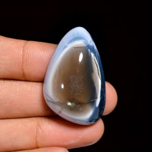 Peut inclure: Une pierre précieuse d'opale en cabochon en forme de larme avec un centre gris foncé entouré d'une couche extérieure blanche et bleue.