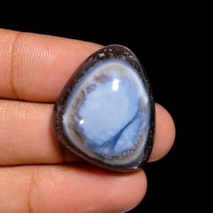 Può includere: Una pietra preziosa di opale nero tagliata a cabochon con un centro blu e bianco e un bordo esterno grigio scuro.