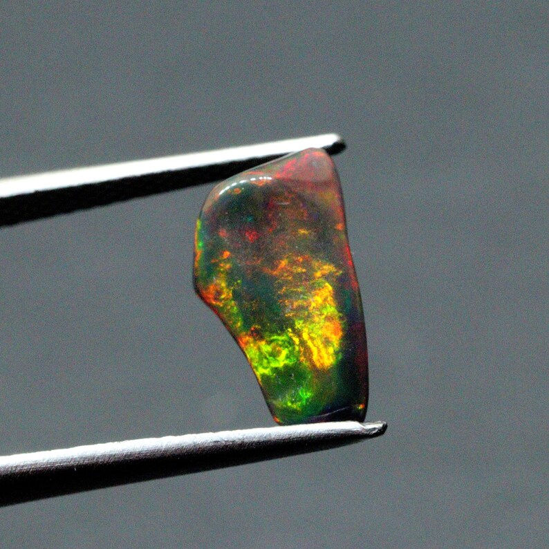 May include: A small, irregularly shaped, dark green opal gemstone with flashes of red, yellow, and green. The opal is held in tweezers.