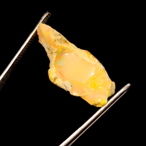 Könnte beinhalten: Ein roher, ungeschliffener Opal-Edelstein, der von einer Pinzette vor einem schwarzen Hintergrund gehalten wird. Der Opal zeigt einen cremefarbenen, durchscheinenden Körper mit gelben und grünen Blitzen und eine raue, strukturierte Oberfläche.