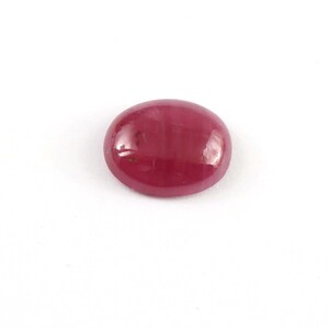 Puede incluir: Una gema de rubí pulida, de forma ovalada. La gema es de color rojo intenso y saturado, con una superficie lisa y reflectante. El rubí se encuentra sobre un fondo blanco liso.