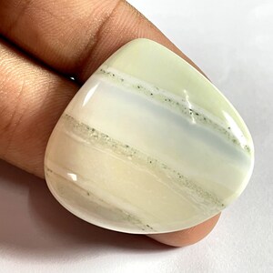 Puede incluir: Un cabujón de ágata blanco y verde con una superficie lisa y pulida. La piedra tiene un patrón natural y orgánico de bandas blancas y verdes.