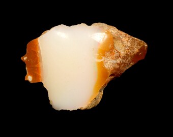 May include: A rough-cut opal gemstone with a creamy white center and a band of orange-brown color along the edges. The stone has a natural, uneven texture and is set against a black background.