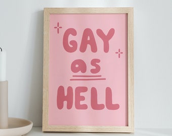 May include: A framed pink print with the text "* GAY as HELL *" in a white, bubbly font. The frame is made of light wood and is sitting on a white surface.
