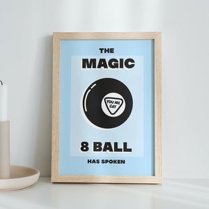 Puede incluir: Una impresión enmarcada con un fondo azul claro que presenta una bola negra de 8 con el texto "You are Gay" dentro del triángulo. El texto "The Magic 8 Ball Has Spoken" está impreso debajo de la bola de 8.