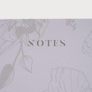 以下が含まれることがあります： 薄い紫色のノート。花柄のデザインとグレーで「NOTES」という文字が印刷されている。