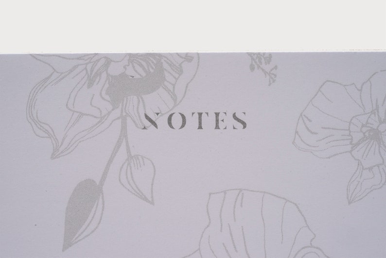 以下が含まれることがあります： 薄い紫色のノート。花柄のデザインとグレーで「NOTES」という文字が印刷されている。