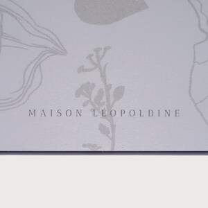 以下が含まれることがあります： 薄いグレーの背景に、花柄のデザインと「MAISON LEOPOLDINE」というテキストが銀色のフォントで書かれています。